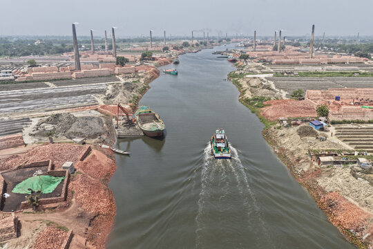 Dhaka, Bangladesh - 12 April 2026: Aerial view of a river flowing through a landscape of industrial brick kilns with tall chimneys and cargo boats navigating the waterway.