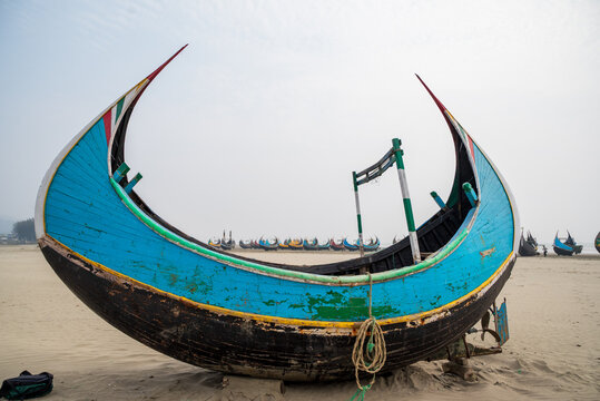 View of a traditional colorful wooden fishing boat resting on the sandy beach with other boats in the distance under a hazy sky in Cox's Bazar, Chittagong Division, Bangladesh.