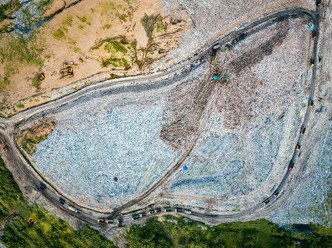 View of a large landfill site with heavy machinery and trucks operating on dirt roads surrounded by vegetation in Denpasar, Bali, Indonesia.