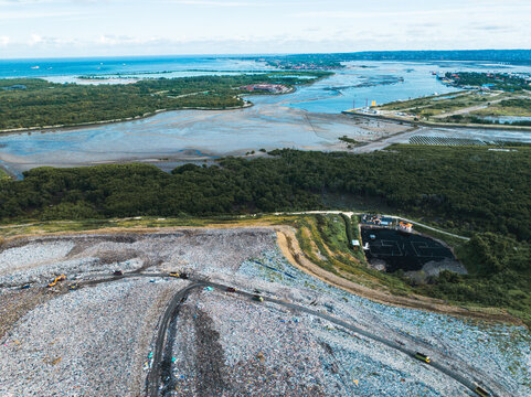 View of a large landfill site with heavy machinery and waste piles situated near lush mangrove forests and the coastline in Denpasar, Bali, Indonesia.