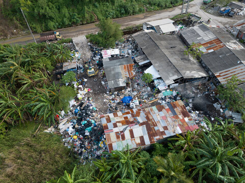 View of a large waste dump and landfill site with piles of plastic trash and debris near industrial sheds surrounded by lush tropical banana trees in Denpasar, Bali, Indonesia.