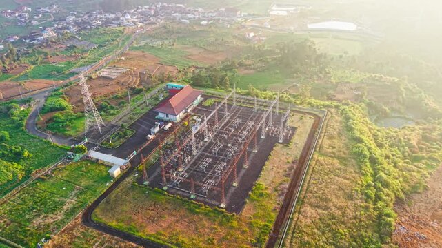 Aerial view of an electrical substation with transmission towers and power lines situated in a rural hilly landscape in Indonesia.