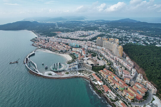 Aerial view of a colorful Mediterranean-style coastal town with a curved pier, sandy beach, and modern buildings nestled between lush green hills and the sea in Phu Quoc, Kien Giang, Vietnam.