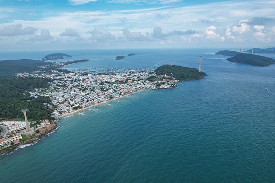Aerial view of a colorful Mediterranean-style coastal town with dense buildings, a sandy beach, and cable car towers along the turquoise sea in Phu Quoc, Kien Giang, Vietnam.