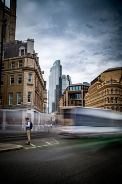 London, United Kingdom - 07 July 2026: View of a man on a street corner in Central London with a blurred bus passing, framed by historic buildings and modern skyscrapers under a cloudy sky.