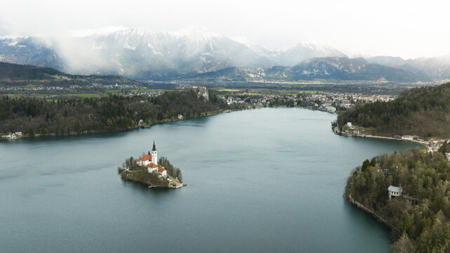 Aerial view of the Pilgrimage Church of the Assumption of Maria on Bled Island, surrounded by the calm waters of Lake Bled and the snow-capped Julian Alps in Bled, Radovljica, Slovenia.