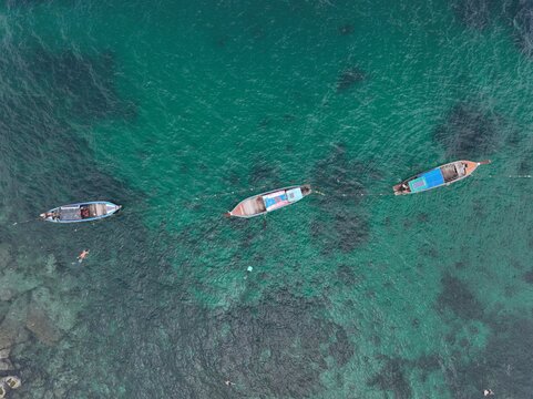 Aerial view of three traditional long-tail boats floating in the clear turquoise waters of the Andaman Sea near Ko Hin Ngam Noi, Ko Tarutao, Satun, Thailand.