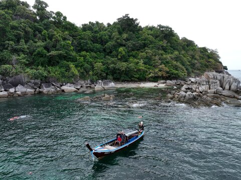 Aerial view of a long-tail boat carrying people near the rocky coastline of Ko Hin Ngam Noi, with lush green forest and clear turquoise water in Ko Tarutao, Satun, Thailand.