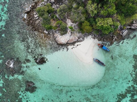 Aerial view of Ko Hin Ngam Noi featuring a white sand beach, turquoise waters, rocky shoreline, lush vegetation, and two long-tail boats anchored in Ko Tarutao, Satun, Thailand.