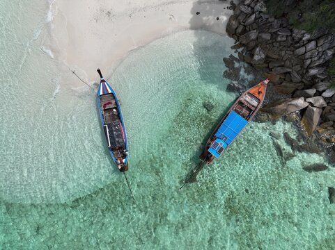 Aerial view of two traditional Thai long-tail boats anchored in clear turquoise water near the sandy shore and rocky coastline of Ko Hin Ngam Noi, Ko Tarutao, Satun, Thailand.