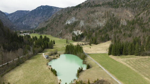 Aerial view of Lake Kreda with its turquoise water surrounded by lush green fields, dense pine forests, and dramatic mountain slopes in Zgornje Gorje, Radovljica, Slovenia.