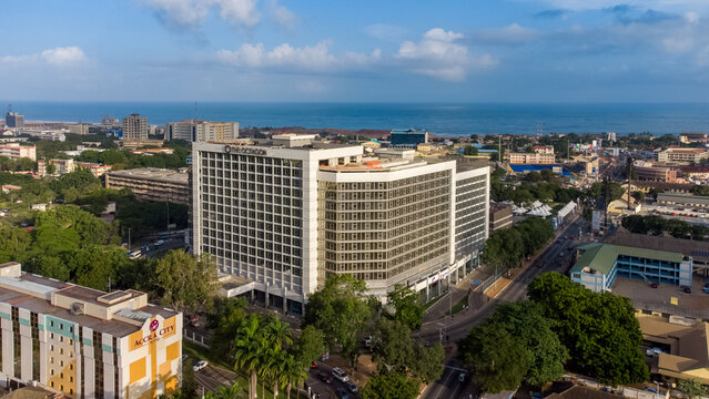 Accra, Ghana - 25 August 2021: Aerial view of The Octagon building in Accra Central, surrounded by urban infrastructure, lush trees, and the coastline in the background under a blue sky.