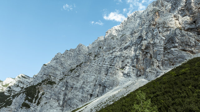 Aerial view of the rugged limestone peaks of Triglav National Park with steep rocky slopes and dense coniferous forests at the base under a clear blue sky in Bohinjsko jezero, Slovenia.