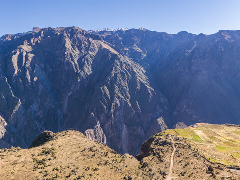 Aerial view of Colca Canyon featuring rugged mountain terrain, deep valley slopes, and terraced agricultural fields under a clear blue sky in Cabanaconde, Arequipa, Peru.
