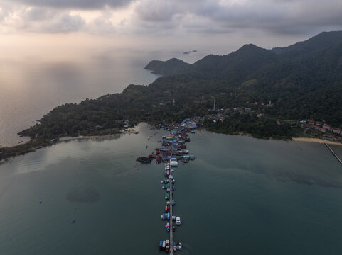 Aerial view of Bang Bao Pier extending into the sea with a traditional fishing village, moored boats, and lush green hills under a soft sunset sky in Koh Chang, Trat, Thailand.