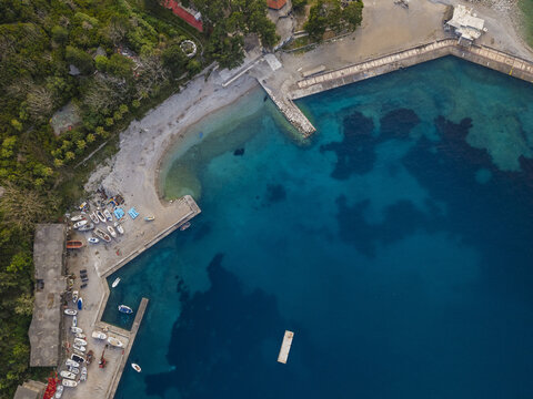 Aerial view of the harbor and coastline in Budva with small boats docked at the pier, turquoise water, and lush greenery in Budva, Budva Municipality, Montenegro.