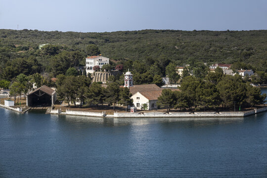 View of the historic Lazareto de Mahon quarantine station surrounded by pine trees and calm blue waters in the port of Mao, Balearic Islands, Spain.