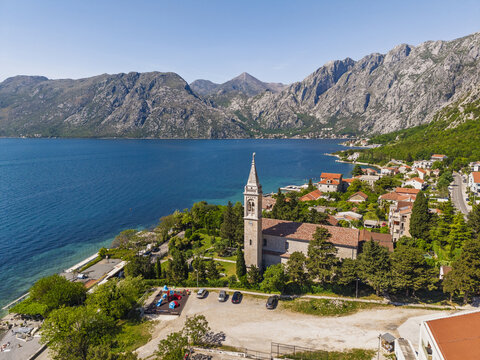 Aerial view of the Church of St. Matthew in Dobrota, with its stone bell tower, trees, terracotta houses, and the blue waters of the Bay of Kotor, Dobrota, Kotor Municipality, Montenegro.
