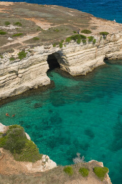 Aerial view of Grotta Sfondata with turquoise water, limestone cliffs, and people swimming along the rocky coastline in Otranto, Puglia, Italy.