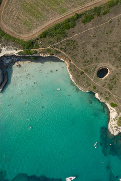 Aerial view of Grotta Sfondata with its distinctive sinkhole on the rocky coastline overlooking a turquoise cove filled with swimmers and small boats in Otranto, Puglia, Italy.