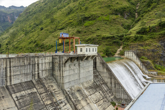 View of the concrete dam structure with water cascading over the spillway, set against lush green mountains along the Ha Giang Loop in Dong Van, Tuyen Quang, Vietnam.