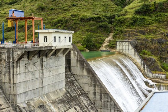 View of the concrete dam structure with water flowing over the spillway, set against lush green hills in the Ha Giang Loop area, Dong Van, Tuyen Quang, Vietnam.