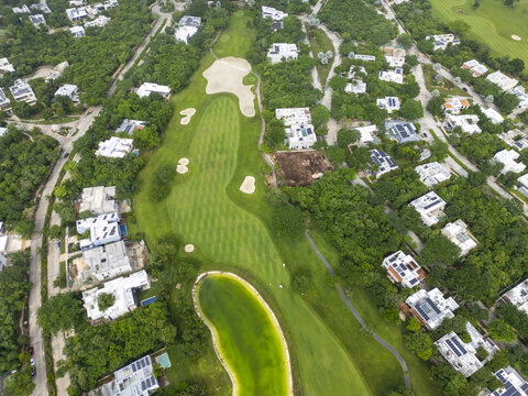 Aerial view of a lush green golf course fairway with sand bunkers and a small pond, surrounded by modern residential houses and dense tropical vegetation in Merida, Yucatan, Mexico.