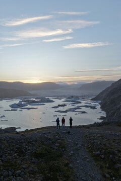 17 August 2025: View of three people standing on a rocky cliff overlooking a glacial lake filled with icebergs under a soft sunset sky.