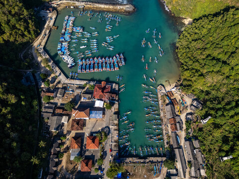 Aerial view of Sadeng Beach fishing port with colorful boats in turquoise water, surrounded by lush green hills and buildings in Kecamatan Girisubo, Daerah Istimewa Yogya, Indonesia.