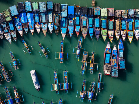 Aerial view of colorful fishing boats moored in the turquoise waters of Sadeng Beach, with wooden hulls and outriggers in Kecamatan Girisubo, Daerah Istimewa Yogya, Indonesia.