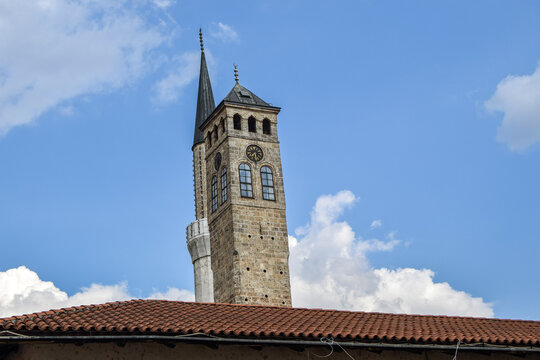 View of the historic Sahat-kula clock tower and a minaret rising above a traditional tiled roof under a bright blue sky in Mostar, Federation of Bosnia, Bosnia and Herzegovina.