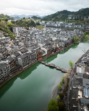 Aerial view of Fenghuang Ancient Town with traditional stilt houses along the Tuojiang River, a stone bridge, and lush mountains under a cloudy sky in Fenghuang, Hunan, China.