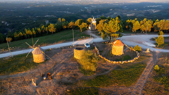 Aerial view of Moinhos de Portela de Oliveira featuring historic stone windmills on a hilltop surrounded by pine forests during golden hour in Sazes do Lorvao, Coimbra, Portugal.