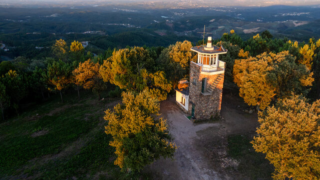 Aerial view of the Moinhos de Portela de Oliveira stone watchtower surrounded by lush forest and rolling hills during golden hour in Sazes do Lorvao, Coimbra, Portugal.