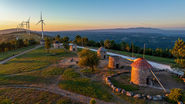 Aerial view of Moinhos de Portela de Oliveira featuring historic stone windmills alongside modern wind turbines on a grassy hilltop in Sazes do Lorvao, Coimbra, Portugal.