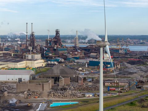 Velsen-Noord, Netherlands - 06 July 2026: Aerial view of Tata Steel plant with a wind turbine in the foreground, industrial infrastructure, smoking chimneys, and piles of raw materials.