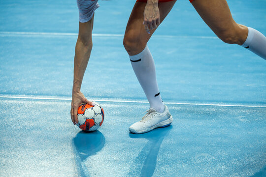Close-up of a handball player's hand touching the ball on the court floor next to white sneakers and socks during a break or preparation for a throw.