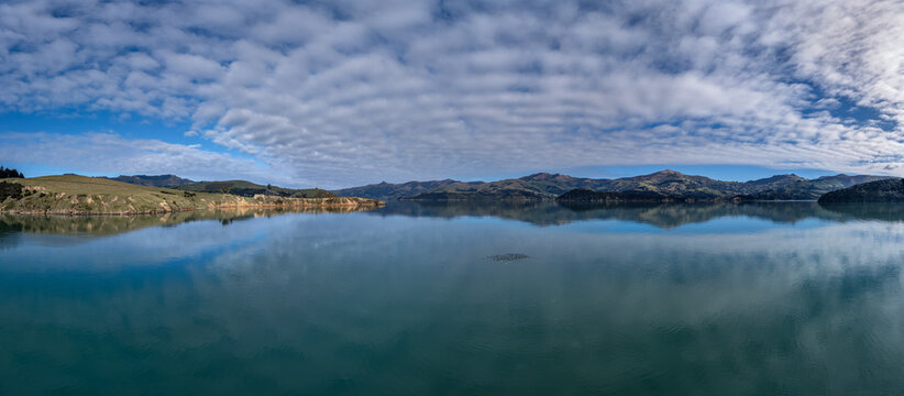 Aerial view of Takamatua Bay featuring calm turquoise waters reflecting rolling hills and a dramatic sky with scattered clouds in the Canterbury Region, New Zealand.