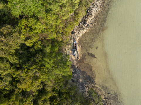 Aerial view of a shipwreck resting on a rocky beach bordered by dense tropical forest and clear coastal waters in Koh Kood, Thailand.