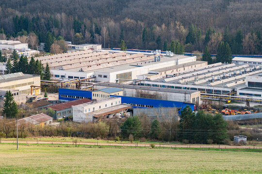 Aerial view of the PPS GROUP a.s. industrial facility with large factory buildings, sawtooth roofs, and blue facade sections, surrounded by forest in Viglas, Banskobystricky kraj, Slovakia.