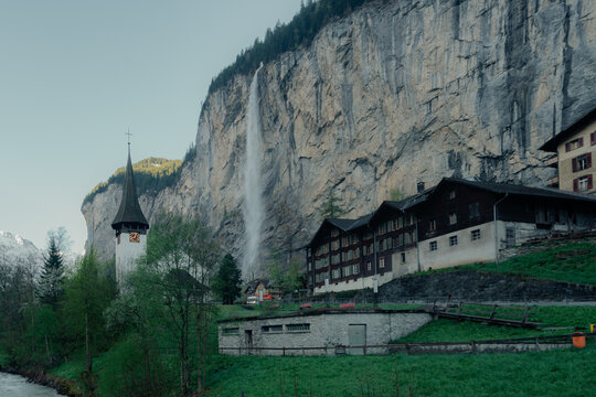 View of the iconic Staubbach Falls cascading down a massive cliff face next to a traditional church and wooden houses in the Lauterbrunnen valley, Switzerland.