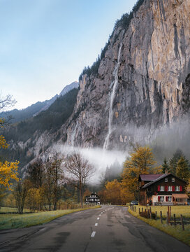 View of the Staubbach Falls cascading down a massive cliff face in the Lauterbrunnen Valley with a traditional wooden house and a winding road in the foreground, Switzerland.