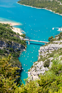 View of Verdon Gorge with turquoise water, a bridge crossing the river, and small boats floating below surrounded by rocky cliffs and lush greenery, Provence-Alpes-Cote d'Azur, France.