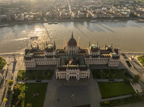 Aerial view of the Hungarian Parliament Building in Budapest, Hungary, featuring its iconic dome, intricate gothic revival architecture, and surrounding gardens along the Danube River at sunset.