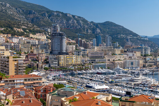 View of the harbor and cityscape of Monaco City with luxury yachts docked at Port Hercules against a backdrop of mountains and clear blue sky in Monaco City, Monaco.