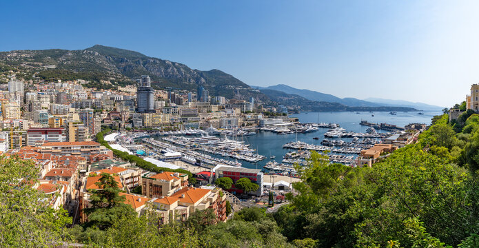 View of Port Hercules in Monaco City with numerous luxury yachts and sailboats docked in the harbor, surrounded by dense urban architecture, hills, and a clear blue sky, Monaco City, Monaco.