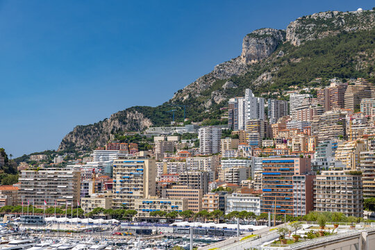 View of the dense urban landscape of Monaco City with high-rise residential buildings, a bustling harbor filled with yachts, and steep mountains in Monaco City, Monaco.