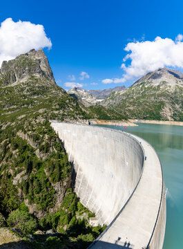 View of the massive concrete arch of the Emosson Dam holding back the turquoise reservoir water, surrounded by steep rocky mountain slopes in Le Chatelard, Valais, Switzerland.