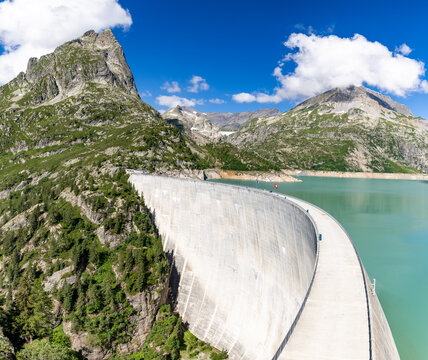 View of the massive concrete wall of the Emosson Dam holding back turquoise reservoir water, surrounded by rugged alpine mountains and green slopes in Le Chatelard, Fribourg, Switzerland.