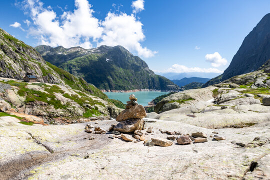 View of VerticAlp Emosson with a stone cairn on rocky terrain overlooking the turquoise Lac d'Emosson and alpine mountains under a blue sky in Le Chatelard, Fribourg, Switzerland.